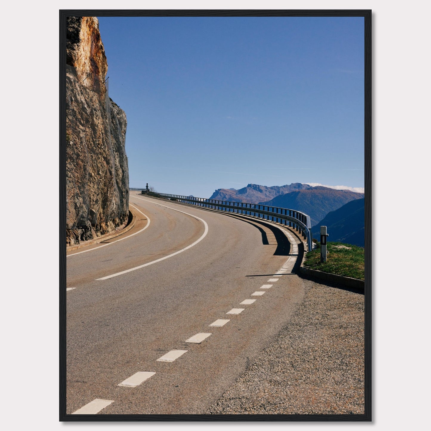 This captivating photograph showcases a winding mountain road bordered by a sheer rock face on one side and a protective guardrail on the other. The clear blue sky and distant mountain range add to the scene's serene beauty.