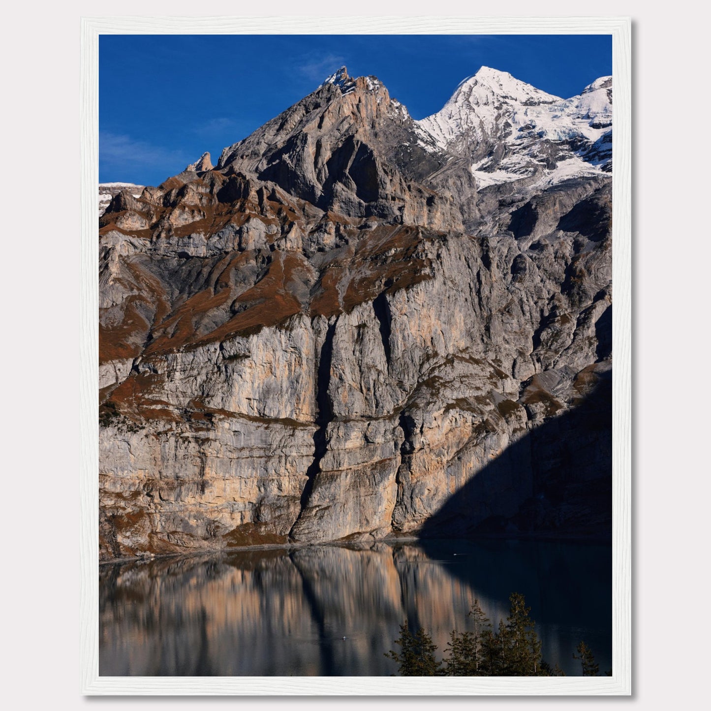 This stunning photograph captures the majestic beauty of a rocky mountain range, with snow-capped peaks under a clear blue sky. The rugged cliffs are reflected in a serene lake below, creating a mirror-like effect. Tall evergreen trees frame the bottom of the scene, adding a touch of greenery to the dramatic landscape.