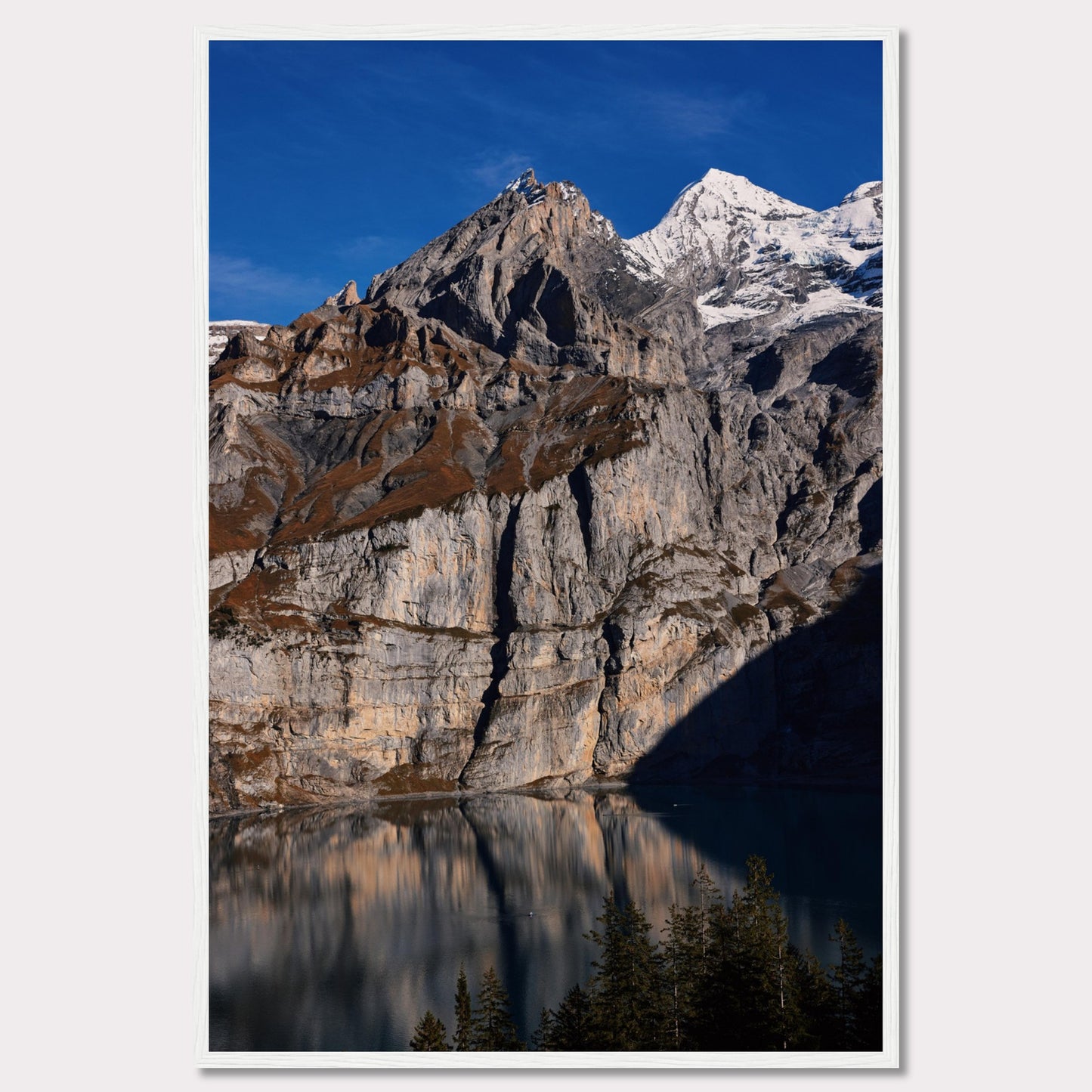 This stunning photograph captures the majestic beauty of a rocky mountain range, with snow-capped peaks under a clear blue sky. The rugged cliffs are reflected in a serene lake below, creating a mirror-like effect. Tall evergreen trees frame the bottom of the scene, adding a touch of greenery to the dramatic landscape.