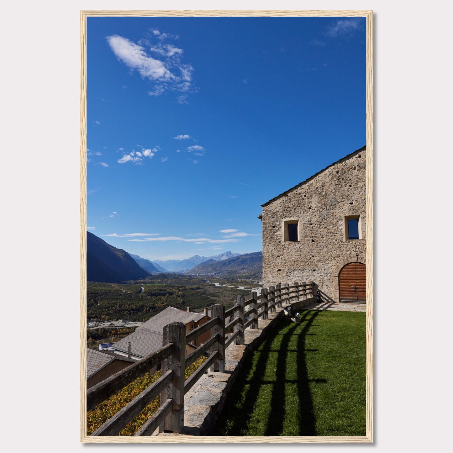 This stunning photograph captures a serene mountain landscape under a clear blue sky. The image features a rustic stone building with wooden windows and an arched wooden door. A wooden fence runs along the green grass, casting shadows that add depth to the scene. In the distance, majestic mountains stretch out, creating a breathtaking view.