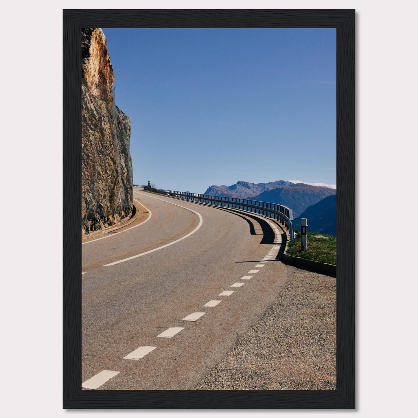 This captivating photograph showcases a winding mountain road bordered by a sheer rock face on one side and a protective guardrail on the other. The clear blue sky and distant mountain range add to the scene's serene beauty.