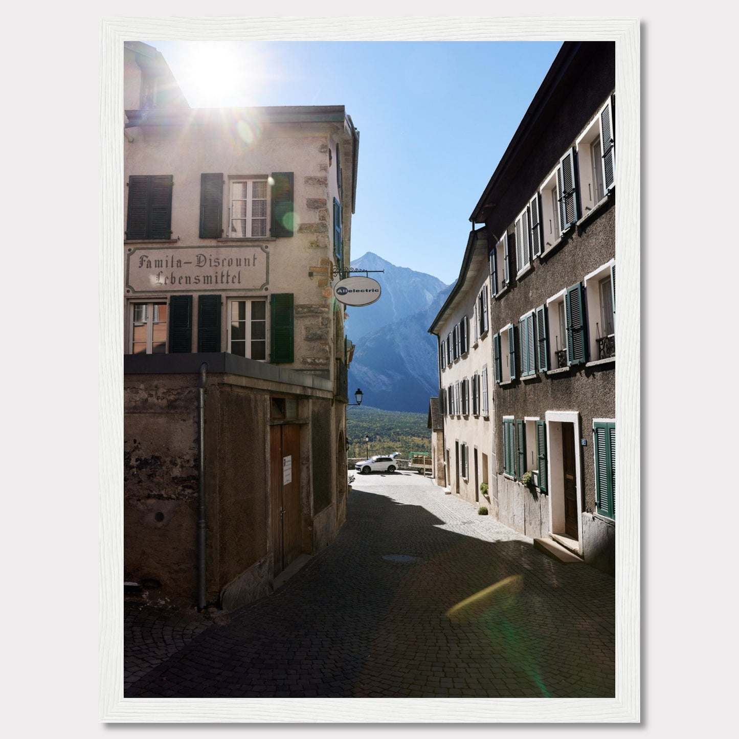 This picturesque scene captures a quaint European street bathed in sunlight, with charming buildings lining the cobblestone path. The sun peeks over the rooftops, casting a warm glow on the surroundings. In the background, majestic mountains rise against a clear blue sky.