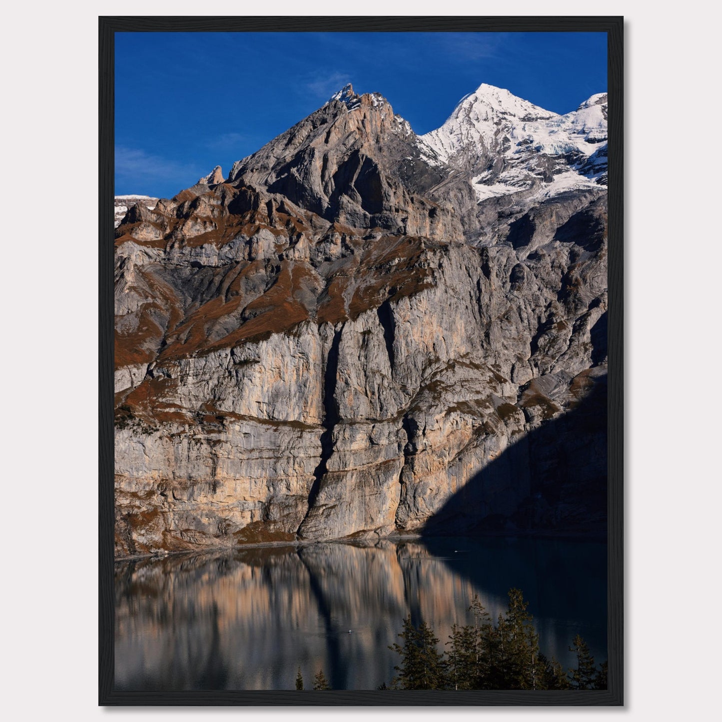 This stunning photograph captures the majestic beauty of a rocky mountain range, with snow-capped peaks under a clear blue sky. The rugged cliffs are reflected in a serene lake below, creating a mirror-like effect. Tall evergreen trees frame the bottom of the scene, adding a touch of greenery to the dramatic landscape.