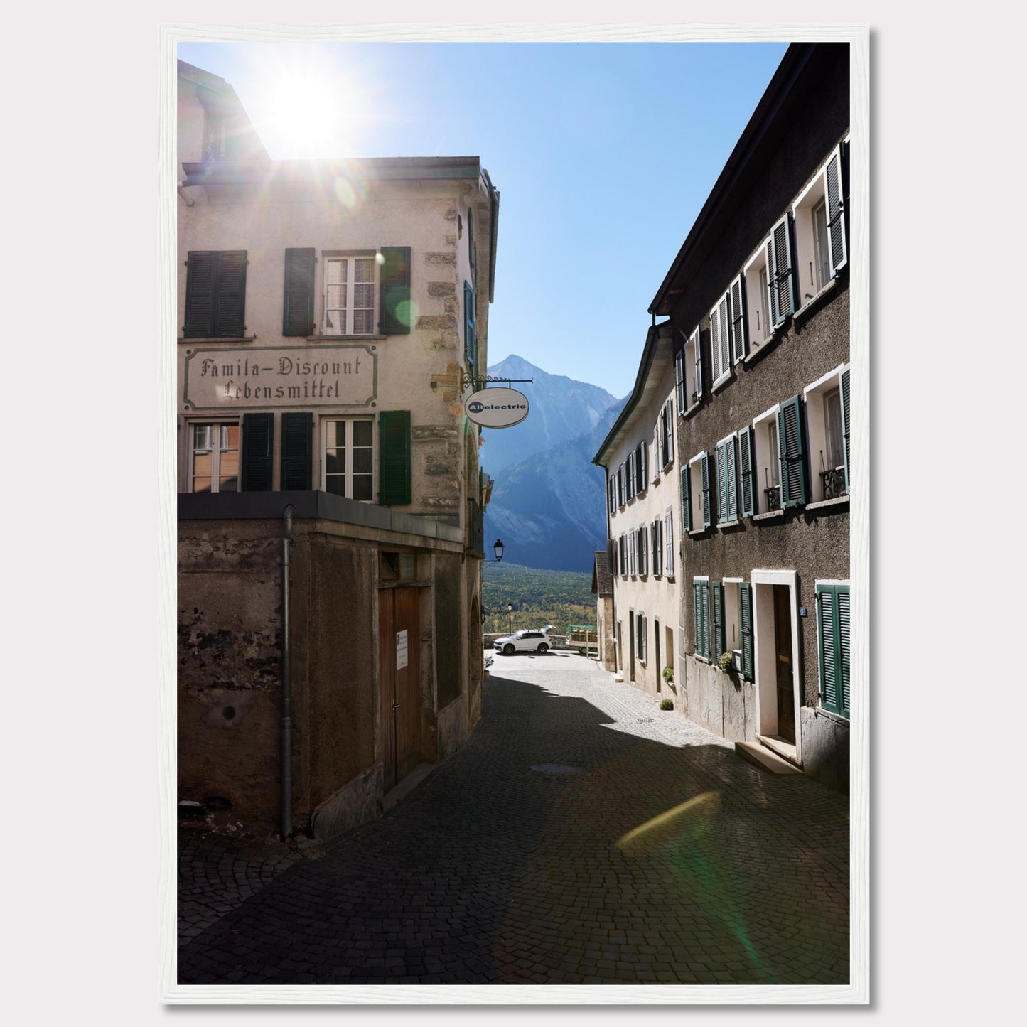 This picturesque scene captures a quaint European street bathed in sunlight, with charming buildings lining the cobblestone path. The sun peeks over the rooftops, casting a warm glow on the surroundings. In the background, majestic mountains rise against a clear blue sky.