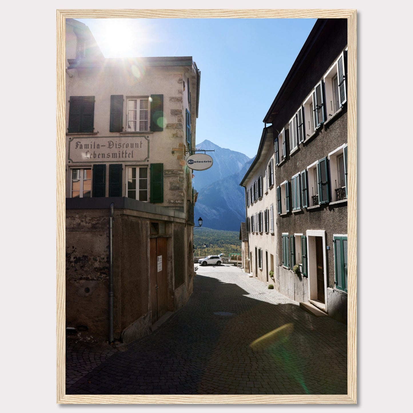 This picturesque scene captures a quaint European street bathed in sunlight, with charming buildings lining the cobblestone path. The sun peeks over the rooftops, casting a warm glow on the surroundings. In the background, majestic mountains rise against a clear blue sky.