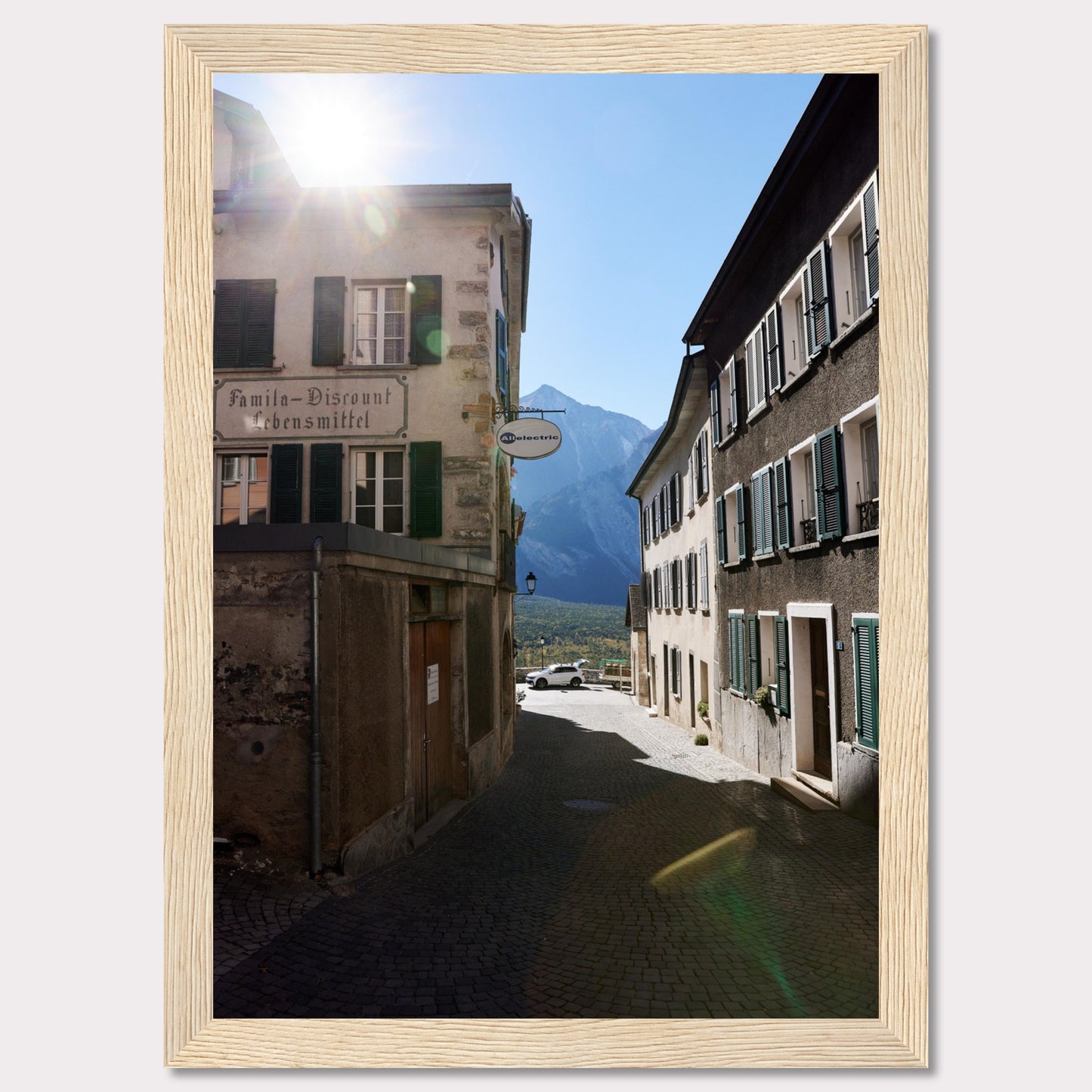 This picturesque scene captures a quaint European street bathed in sunlight, with charming buildings lining the cobblestone path. The sun peeks over the rooftops, casting a warm glow on the surroundings. In the background, majestic mountains rise against a clear blue sky.