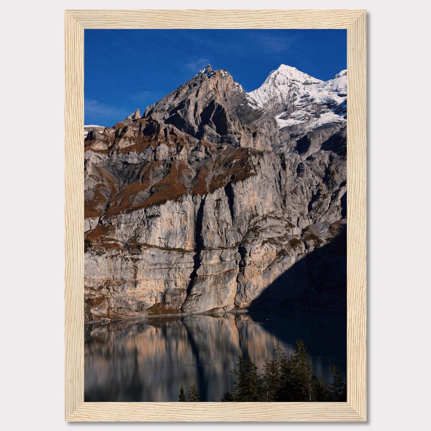 This stunning photograph captures the majestic beauty of a rocky mountain range, with snow-capped peaks under a clear blue sky. The rugged cliffs are reflected in a serene lake below, creating a mirror-like effect. Tall evergreen trees frame the bottom of the scene, adding a touch of greenery to the dramatic landscape.