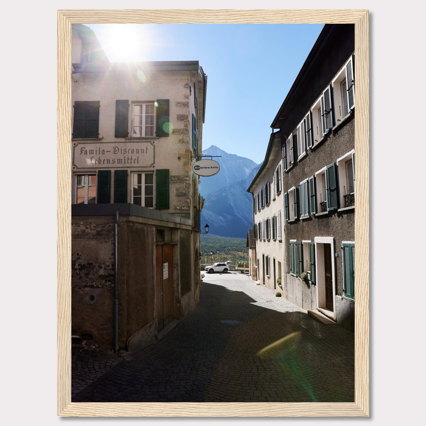 This picturesque scene captures a quaint European street bathed in sunlight, with charming buildings lining the cobblestone path. The sun peeks over the rooftops, casting a warm glow on the surroundings. In the background, majestic mountains rise against a clear blue sky.