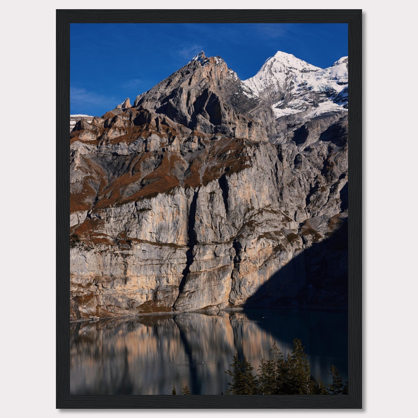 This stunning photograph captures the majestic beauty of a rocky mountain range, with snow-capped peaks under a clear blue sky. The rugged cliffs are reflected in a serene lake below, creating a mirror-like effect. Tall evergreen trees frame the bottom of the scene, adding a touch of greenery to the dramatic landscape.